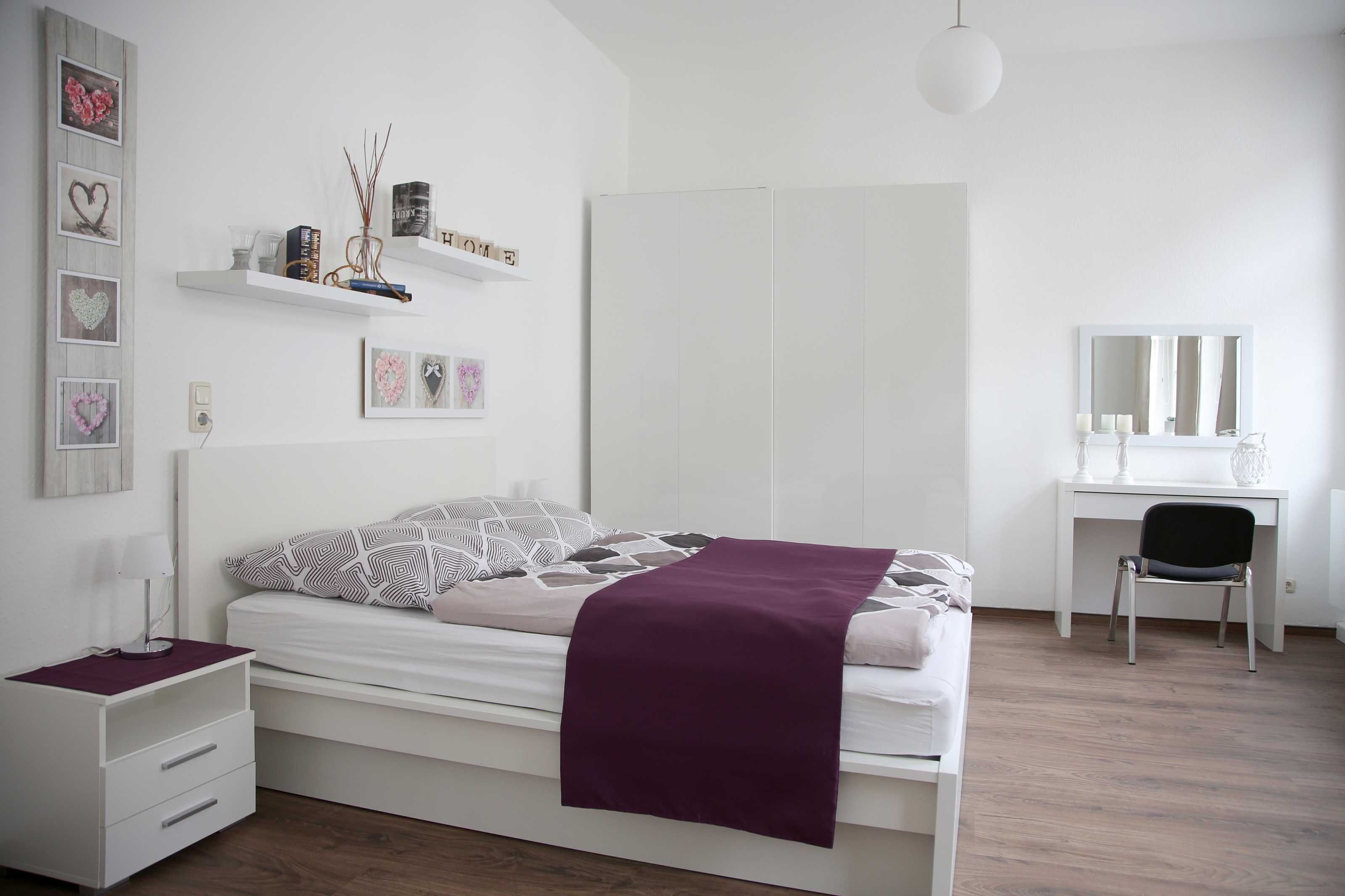 Ein Schlafzimmer mit einem Bett, Nachttisch, Schminktisch und Schreibtisch mit Stuhl. Regale mit Dekorationselementen und ein Spiegel an der Wand sind ebenfalls vorhanden.