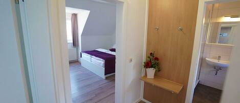 Ein Schlafzimmer mit Bett, einer Holztür, einer kleinen Ablage mit Blumen sowie einem Badezimmer mit Waschbecken und Spiegel.
