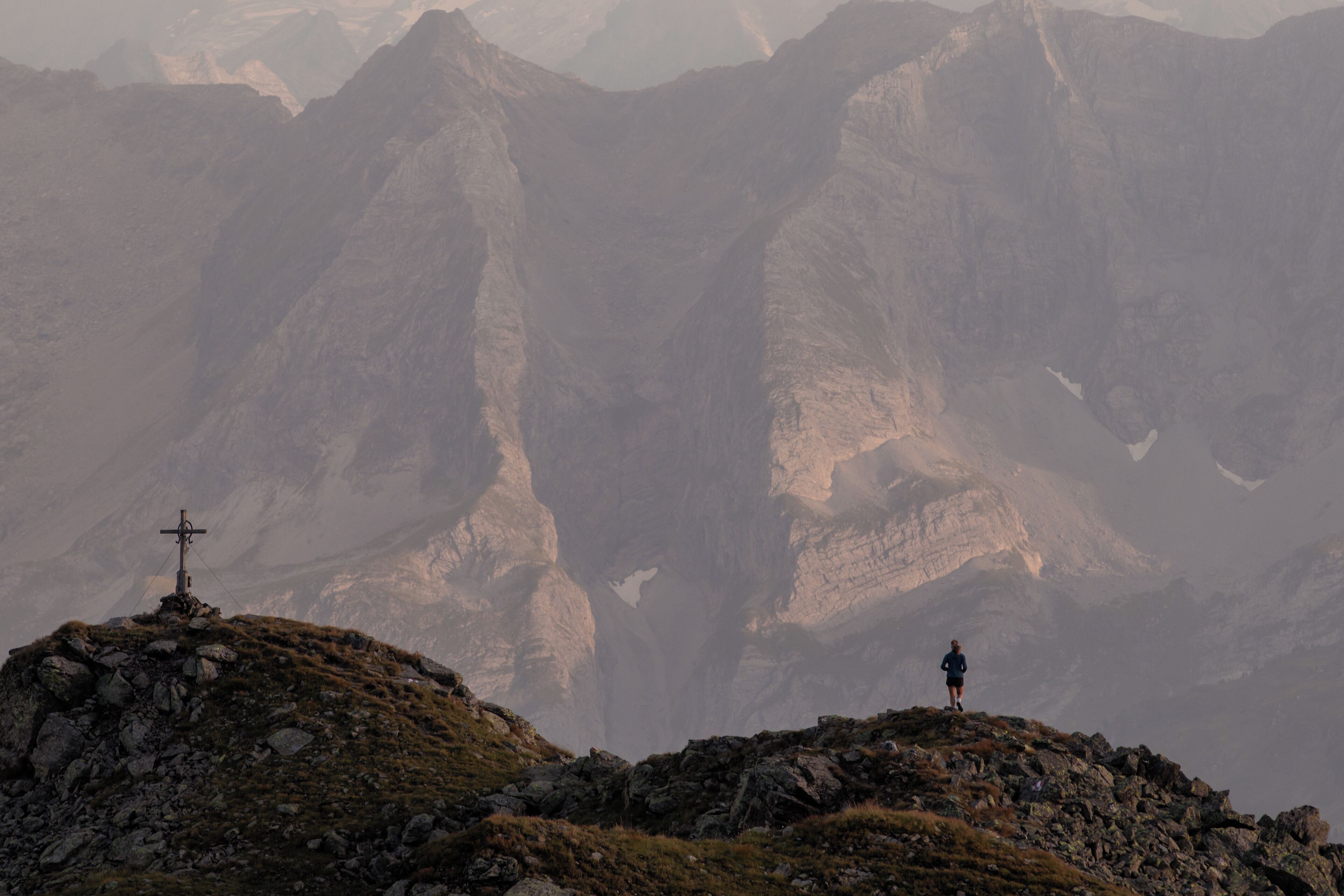 A mountainous landscape with a cross on a rocky outcrop and a person standing on the peak.