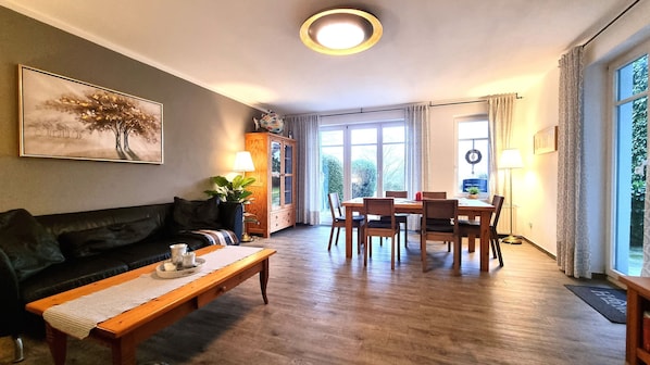 Uma sala de estar com piso de madeira, um sofá, uma mesa de jantar com cadeiras, um armário e uma janela grande com cortinas.