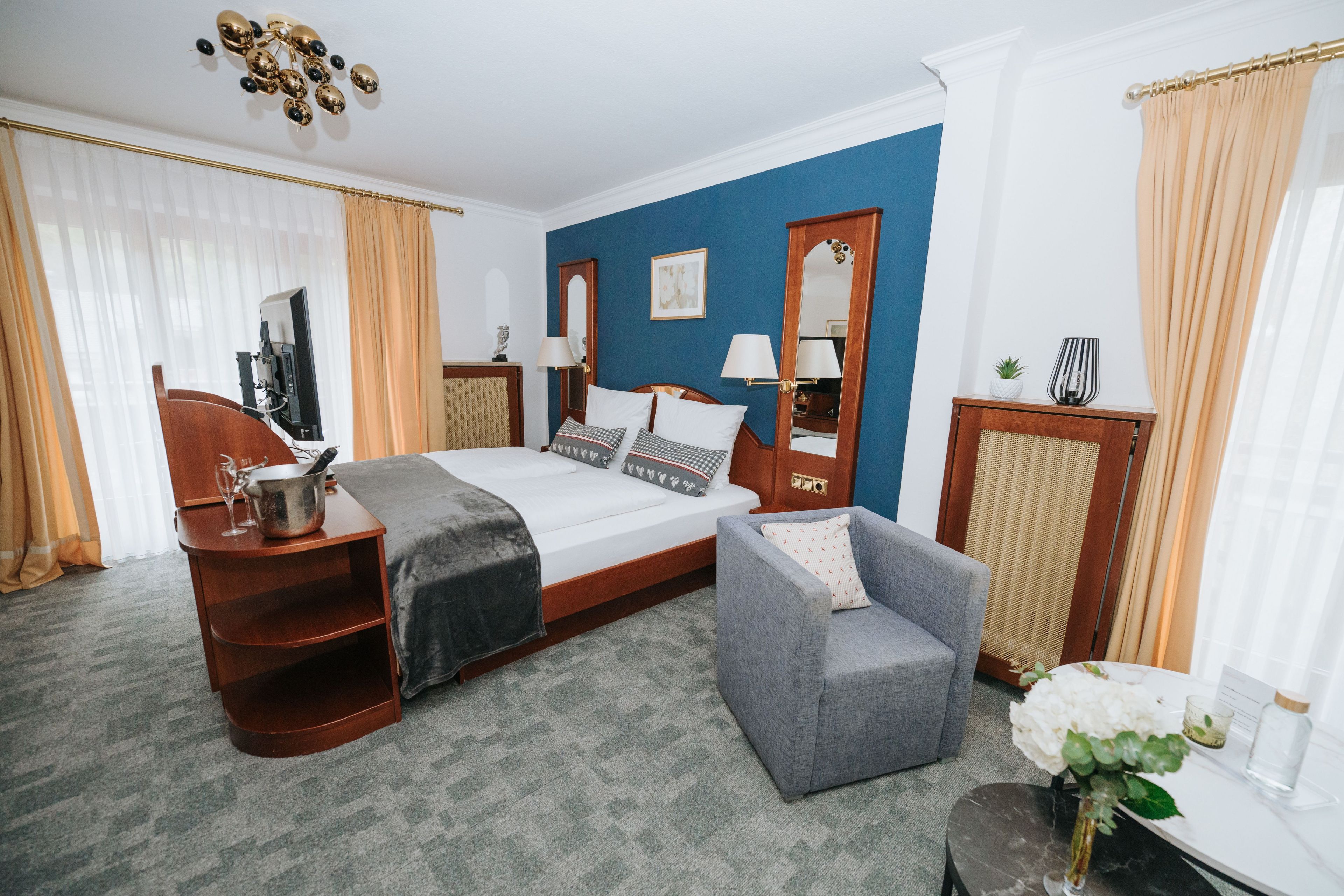 A hotel room with a bed, a desk with a computer, a chair, a small table with a vase, and a mirror.