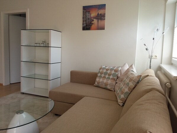 Een woonkamer met een beige bank, gestreepte kussens, een glazen salontafel en een wit wandrek.