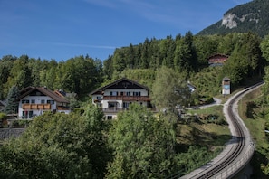 Un hotel en la ladera de una montaña con varias construcciones, rodeado de un bosque denso y una vía de tren.