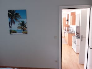 A room with a painting of a beach scene, a door leading to a kitchen with modern appliances, and a wooden floor.