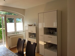 Een eetruimte met een tafel, stoelen, een kast met glazen deuren en een raam met jaloezieën.
