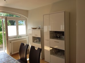 Un comedor con una mesa, sillas, un armario con puertas de cristal y una ventana con persianas.