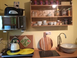 A kitchen with a microwave, kettle, and a sink. There are shelves with dishes and a coffee maker.