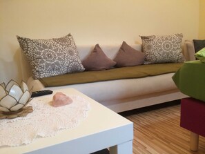 A living room with a white sofa, patterned cushions, a small white table with a decorative object, and a wooden floor.