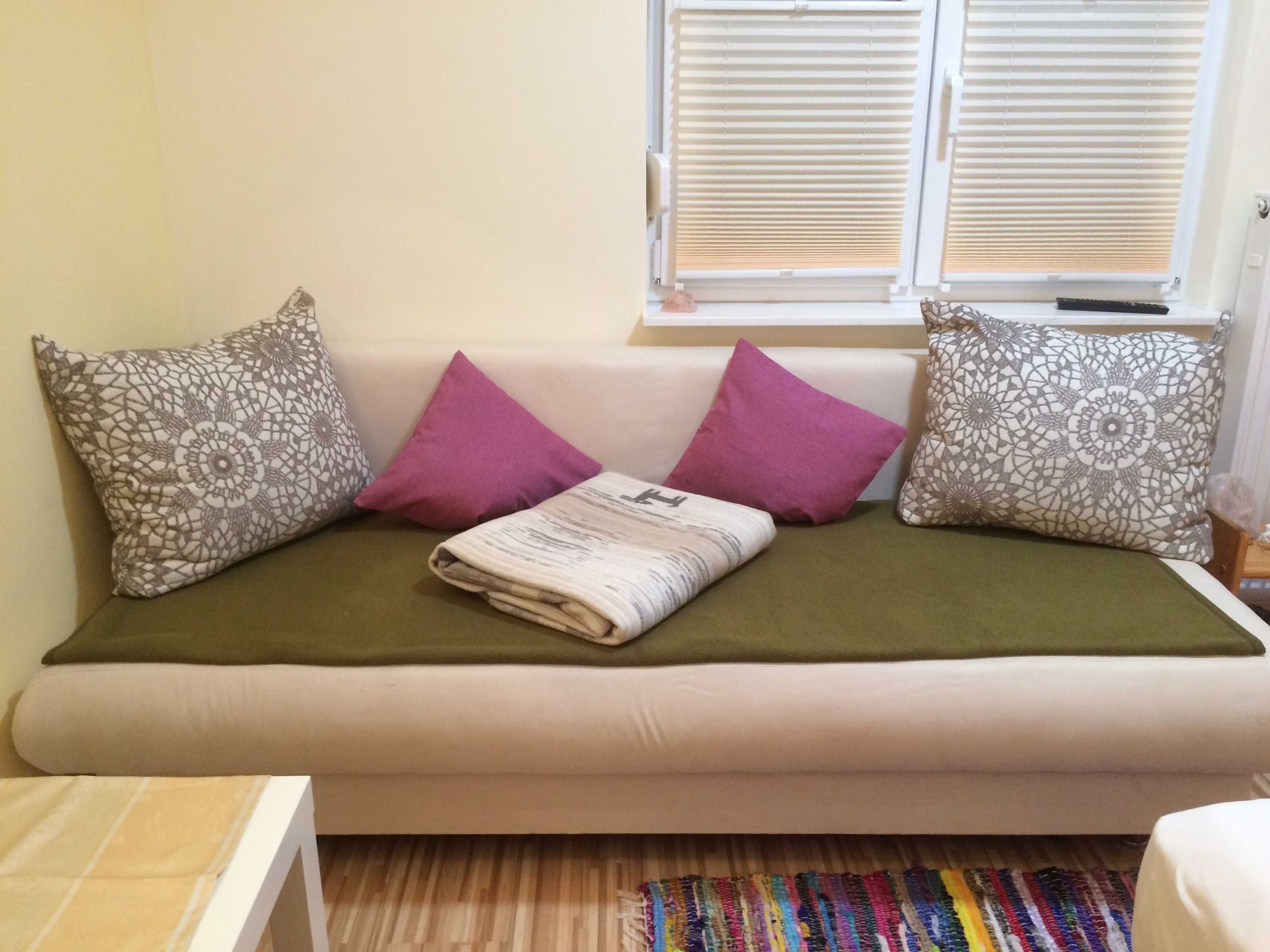 A sofa with patterned cushions, a folded blanket, and a window with blinds.