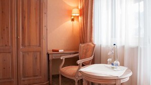 Una habitación de hotel con un armario de madera, un escritorio con silla, una mesita de noche con lámpara y una ventana con cortinas traslúcidas.