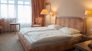 Una habitación de hotel con una cama grande, dos mesitas de noche, una silla, una mesita pequeña y un radiador.