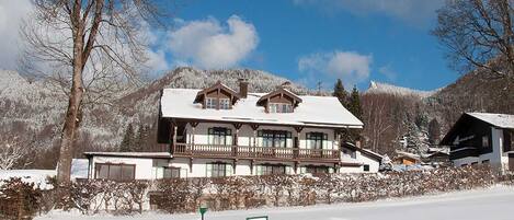 A large, traditional chalet-style building with a snow-covered roof, surrounded by a snowy landscape and mountains.