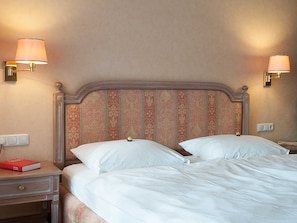A neatly made bed with white linens, a wooden headboard with a patterned design, two bedside lamps, and a nightstand with a book and a lamp.