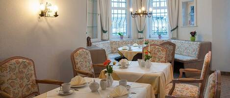 Un coin repas avec des tables dressées pour quatre personnes, des chaises recouvertes d’un tissu à motifs floraux, un lustre et une fenêtre agrémentée de rideaux.