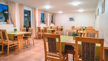 A dining area with multiple wooden tables and chairs, a green tablecloth, and a potted plant on each table.