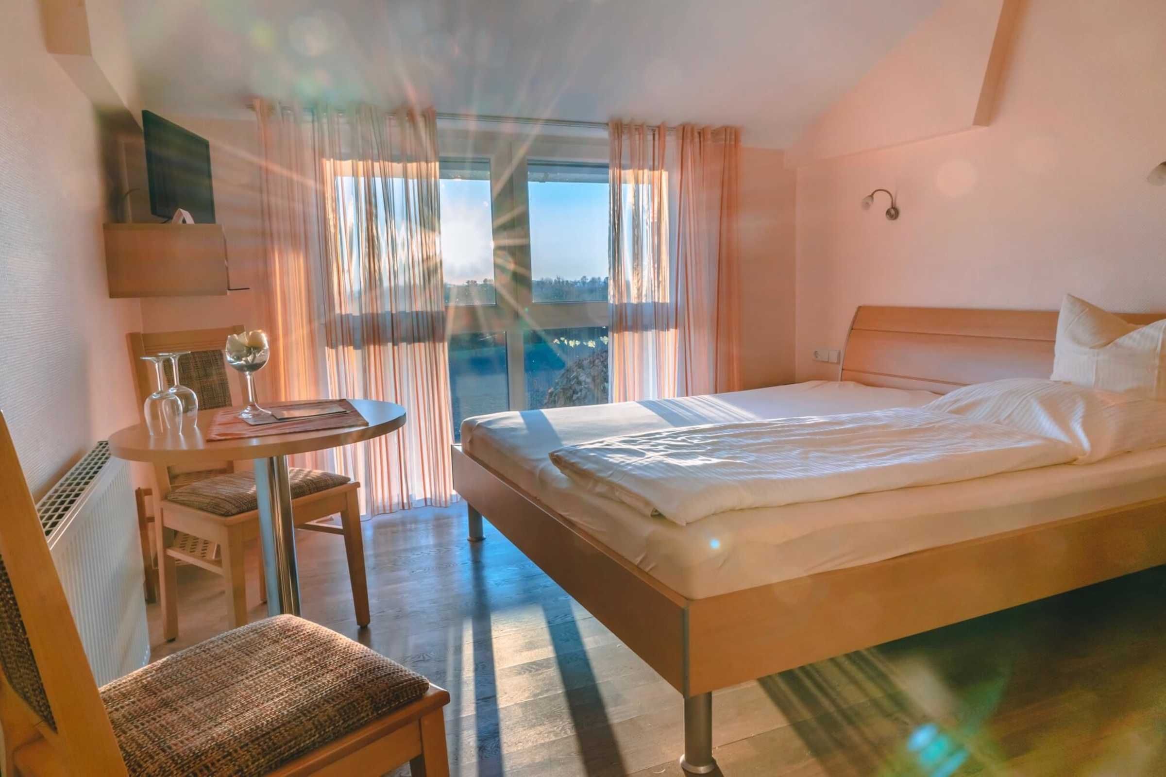 A hotel room with a bed, a small table with chairs, a TV, and a view through large windows.
