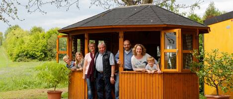 Un kiosque en bois doté d’un toit incliné, entouré de verdure et bordé d’un bâtiment jaune.