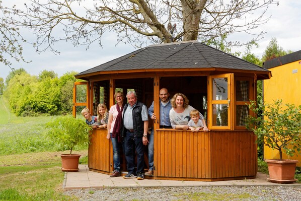 Ein hölzerner Pavillon mit geneigtem Dach, umgeben von Grünflächen und einem gelben Gebäude.
