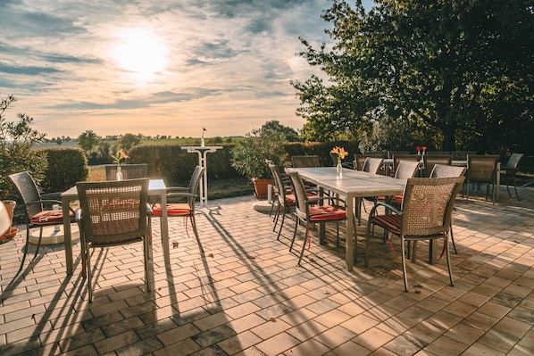 An outdoor patio with a table and chairs, surrounded by greenery and a clear sky.