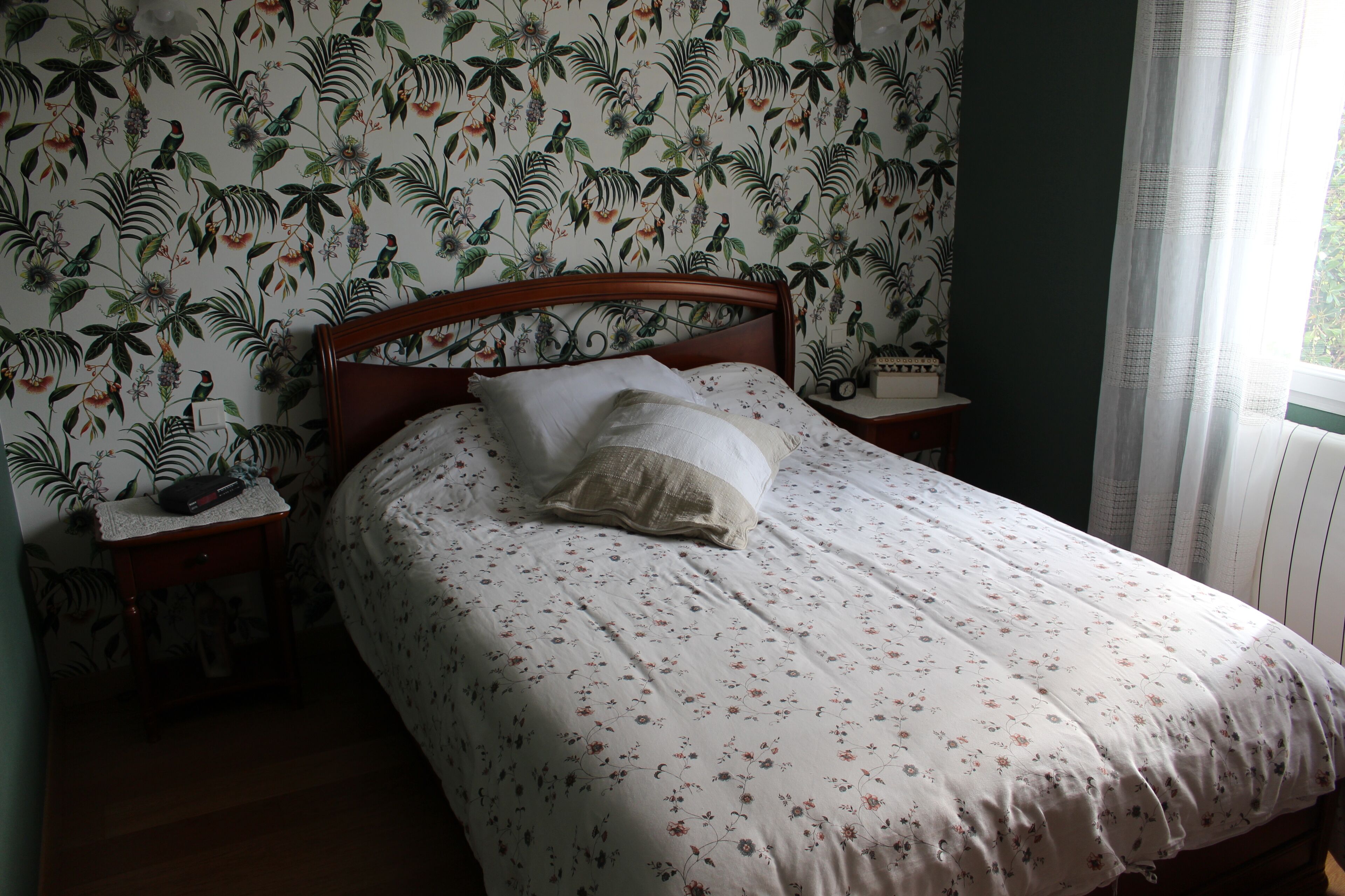Quarto com papel de parede estampado com flores, cabeceira de madeira, colcha branca e mesa de cabeceira com abajur.