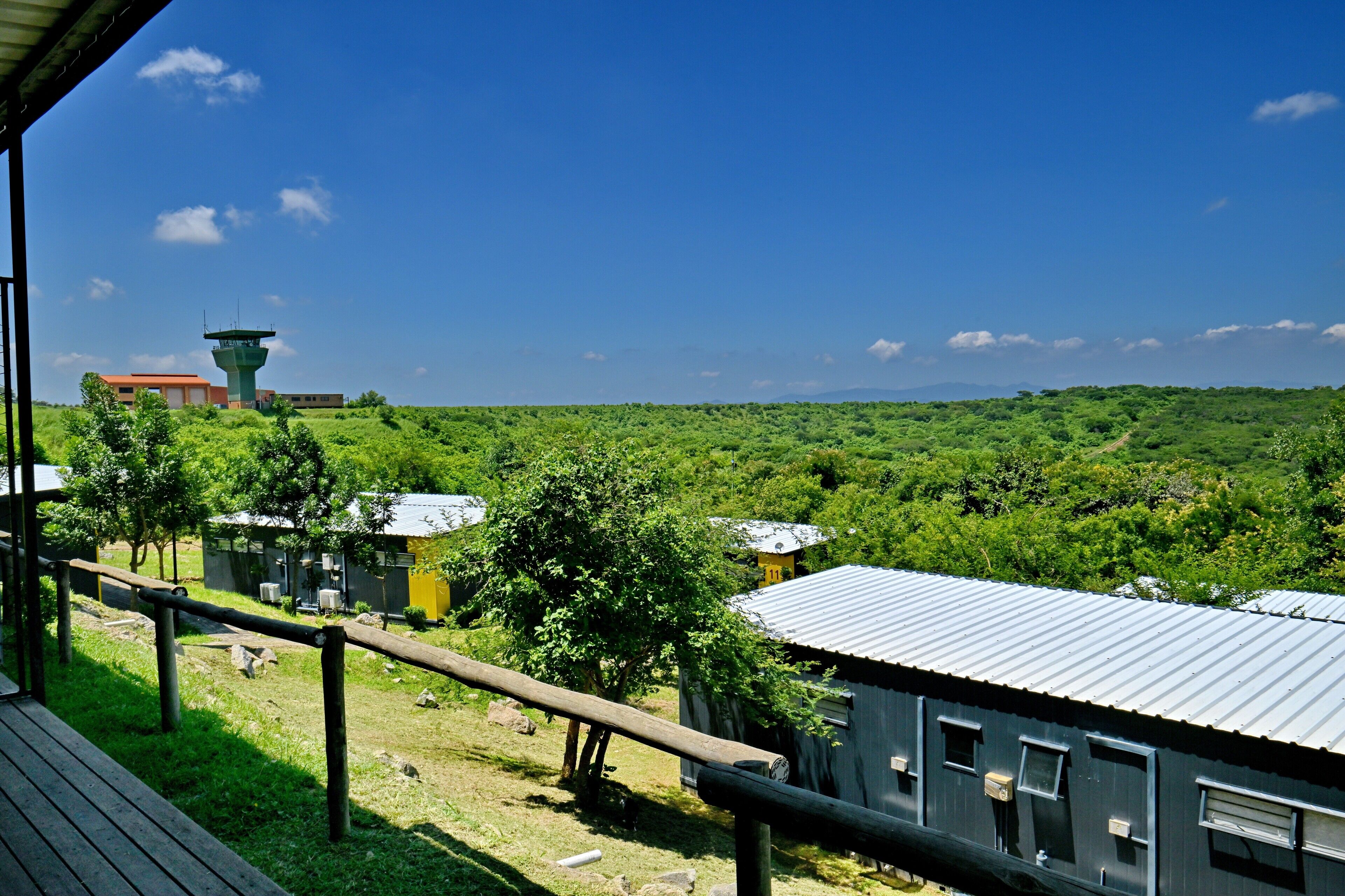 Vista de un paisaje verde con una torre de control, un edificio amarillo y un edificio azul con techo metálico.