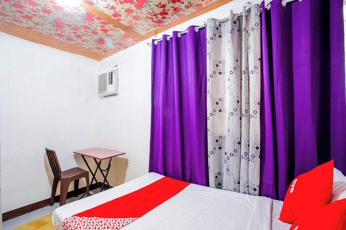 A bedroom with a bed, a small table, a chair, purple curtains, and a floral ceiling.