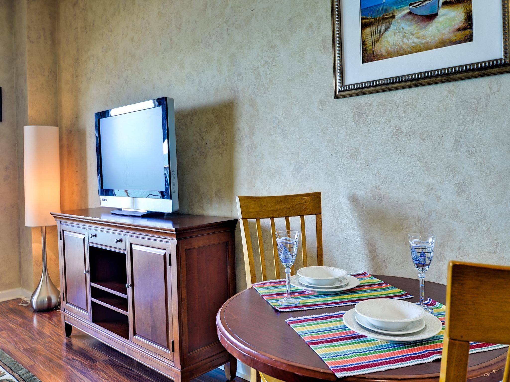 A dining area with a wooden table set for two, a television on a cabinet, and a painting on the wall.