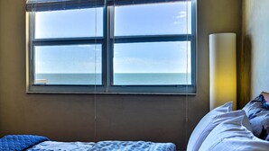 A room with a bed, a window with a view of the sea, and a lamp on the wall. - Regatta Beach Club C713 '90 day min stay with 2 week lead time' (Clearwater Beach)