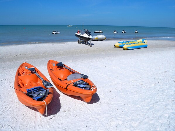 Zwei orangefarbene Kajaks am sandigen Strand, im Hintergrund ein klarer, blauer Himmel und ruhiges Meer.
