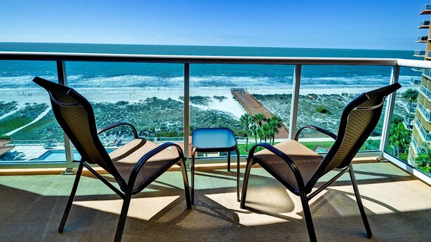 A balcony with two wicker chairs and a small table, overlooking a beach with a pier and ocean waves. - Regatta Beach Club C713 '90 day min stay with 2 week lead time' (Clearwater Beach)