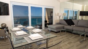 A modern living room with a glass dining table set for four, a grey sofa, and a balcony with a view. - Regatta Beach Club C1009 '90 day min stay with 2 week lead time' (Clearwater Beach)