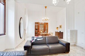 Een woonkamer met een bank, een eettafel met stoelen, een kroonluchter, een spiegel en een radiator.