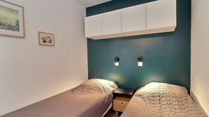 A small, modern bedroom with two single beds, a nightstand, and a wall-mounted cabinet.