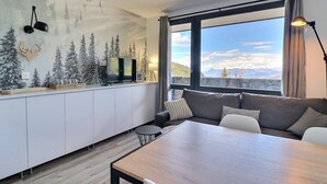 A modern living room with a large window, a sofa, a dining table, and a TV on a white cabinet.