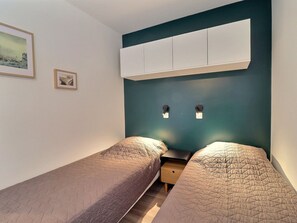 A small, modern bedroom with two single beds, a nightstand, and a wall-mounted cabinet.