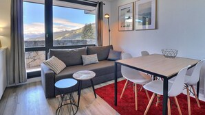 A modern living room with a large window, a sofa, a small round table, and a dining table with chairs.