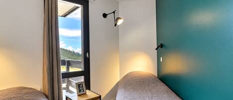 Un dormitorio con dos camas, una mesita de noche con un marco de fotos, una ventana con cortinas y una lámpara de pared.