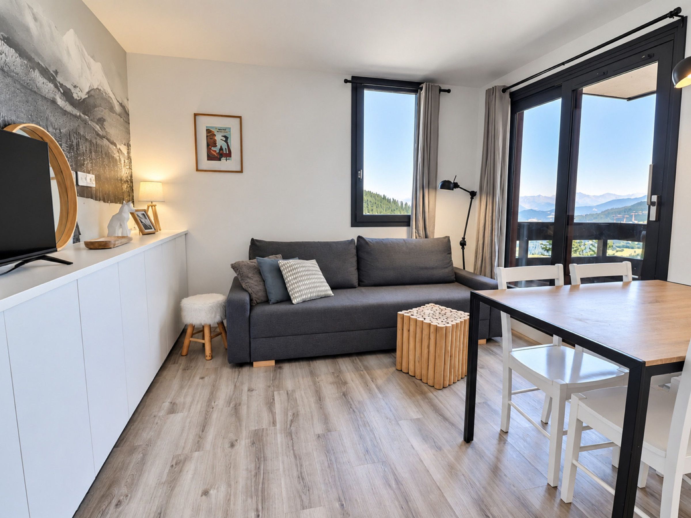 Un salon moderne avec un canapé gris, une table basse en bois, une table à manger avec des chaises et de grandes fenêtres donnant sur l’extérieur.