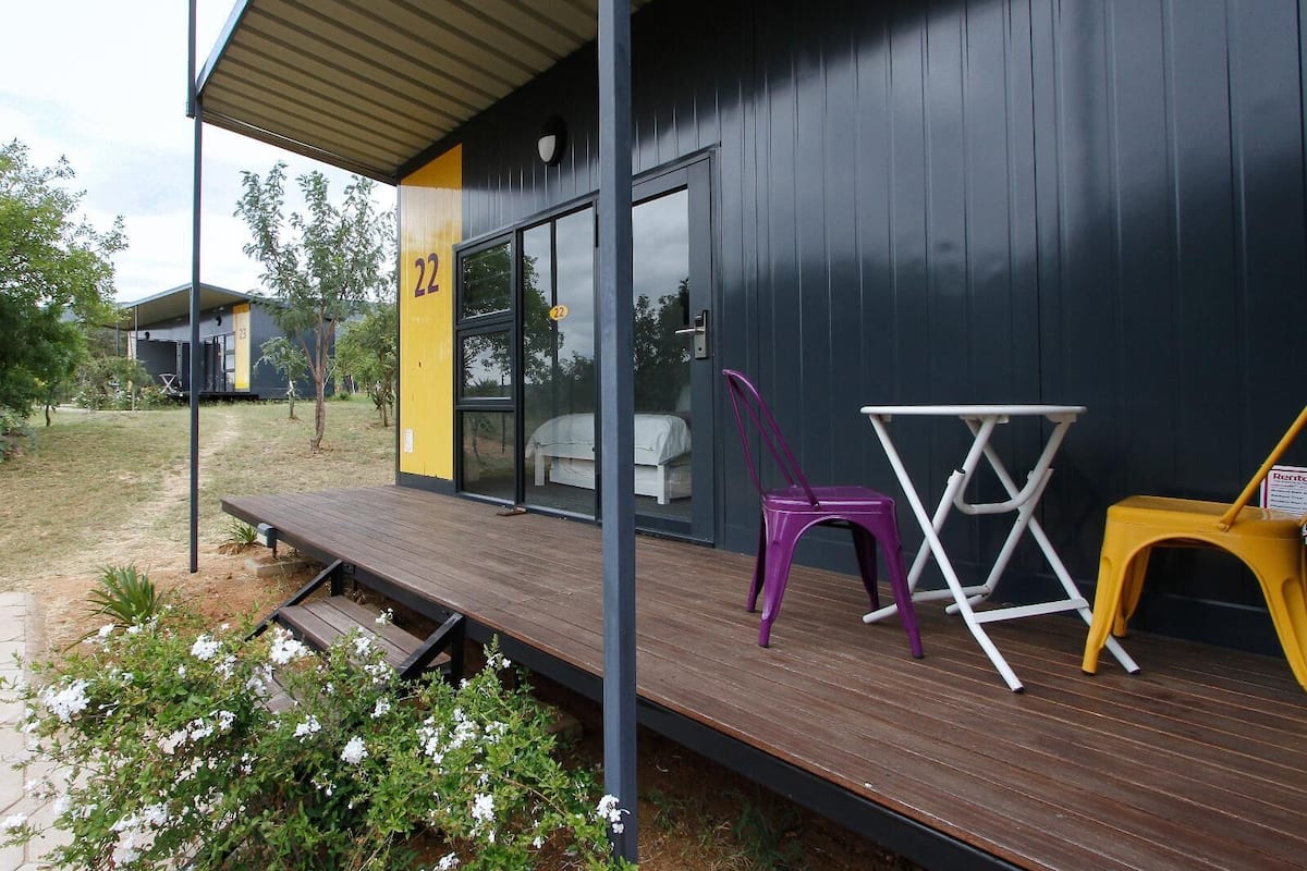 Un edificio moderno con fachada oscura, el número '22' en el lateral, y un espacio al aire libre cubierto con una terraza de madera, una silla morada y una silla amarilla.