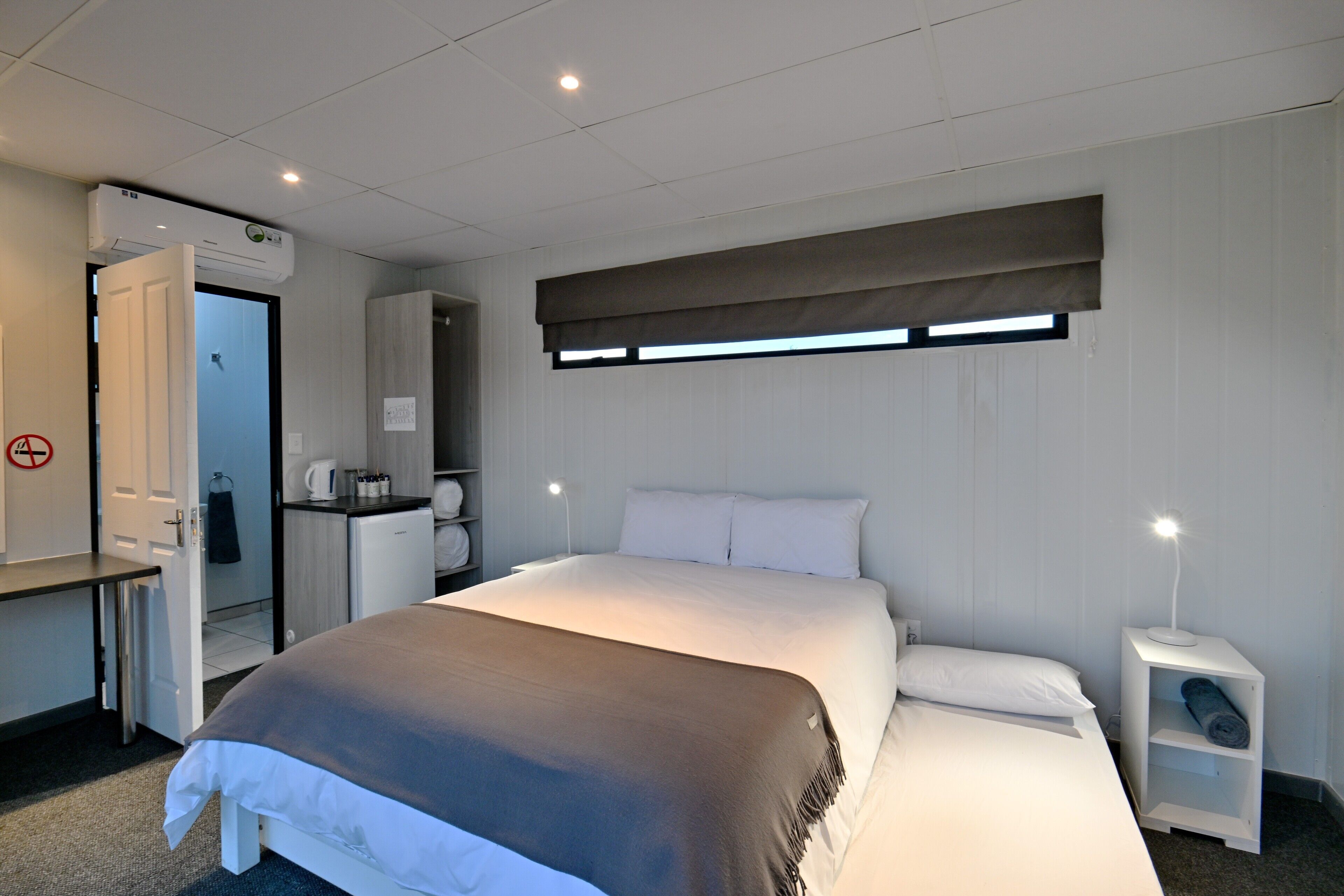 Une chambre d’hôtel comprenant un lit, une table de chevet, une commode et une fenêtre avec des stores.