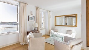 Een ruime kamer met een groot raam en uitzicht op zee, een witte bank, twee rieten fauteuils, een glazen salontafel en een houten kast.