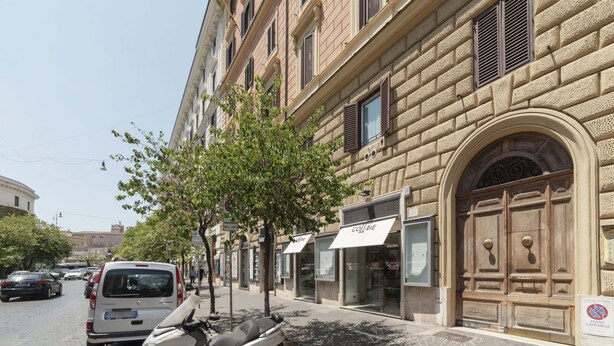 Calle con autos estacionados y una moto, un edificio con una puerta de madera grande, y un cartel que dice 'COLME'.