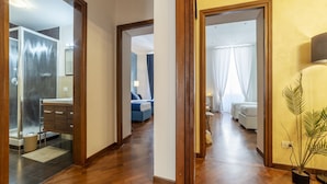 Un pasillo con piso de madera, un baño con ducha de vidrio y un dormitorio con cama y cortinas.