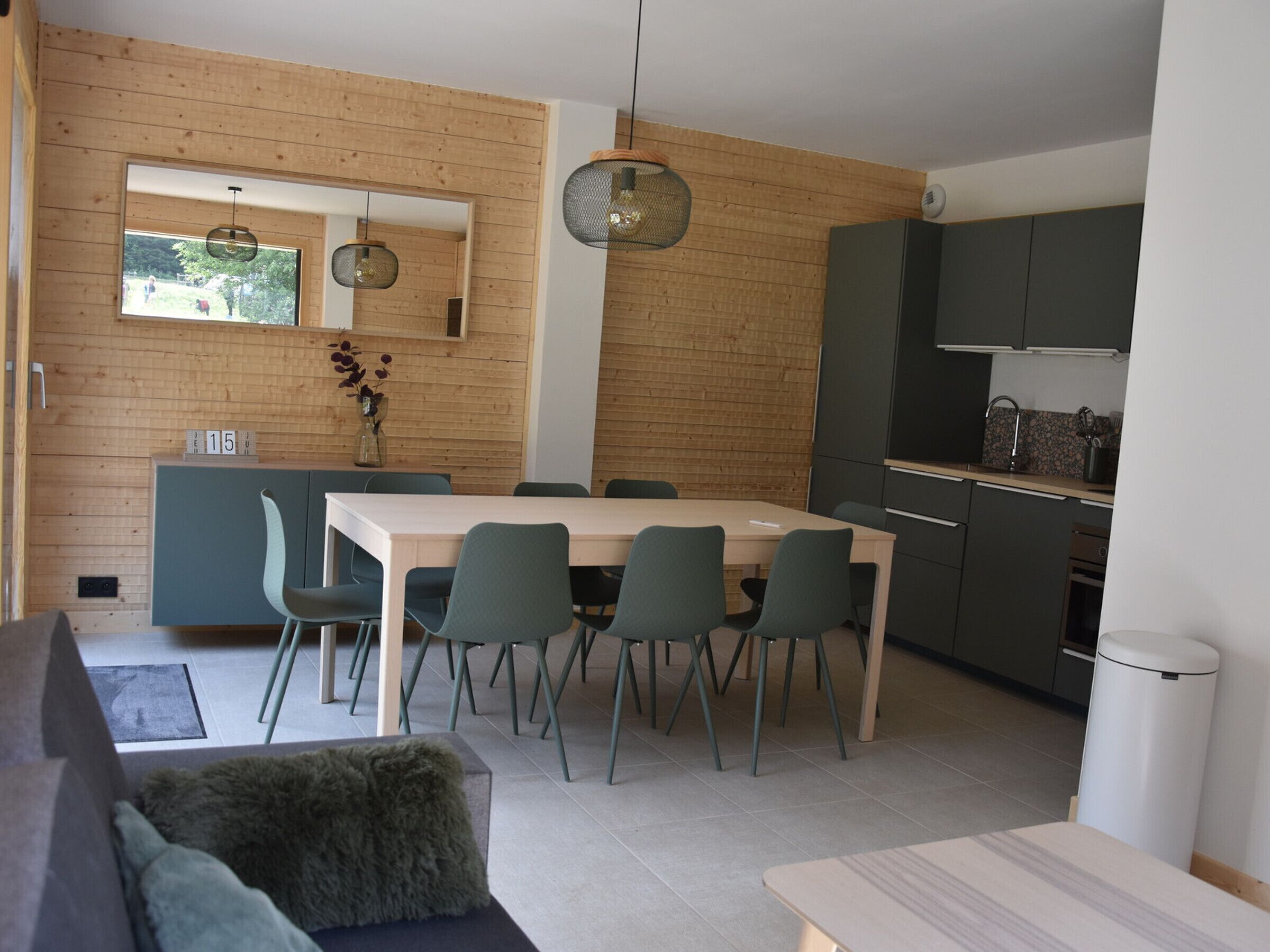 A modern kitchen with dark cabinets, a dining table with chairs, and a wooden wall with a window.