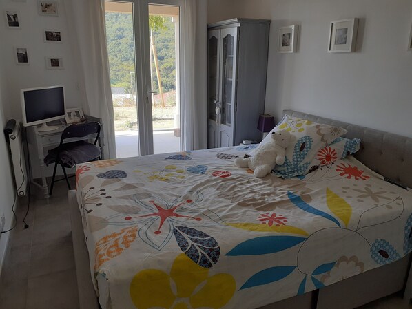 A bedroom with a bed, a desk with a computer, a wardrobe, and a window with a view of greenery.
