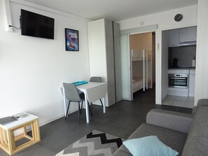 Uma sala de estar moderna com um sofá cinzento, uma mesa de jantar com cadeiras, uma TV na parede e uma área de cozinha com fogão e forno.