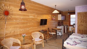 Acogedora habitación de cabaña de troncos con cama, dos sillones de mimbre, una mesita con un florero, televisión y una zona de cocina.