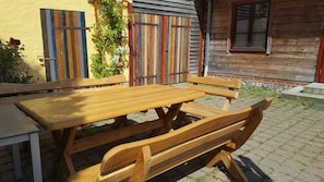 An outdoor wooden table and benches in a garden area with a yellow wall and wooden fence in the background.