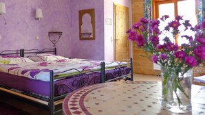 A bedroom with a bed, a round table, a vase with flowers, a wooden door, and a window with curtains.