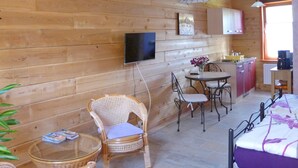 A cozy room with wooden walls, a bed, a dining table with chairs, a TV mounted on the wall, and a kitchen area with cabinets and a window.